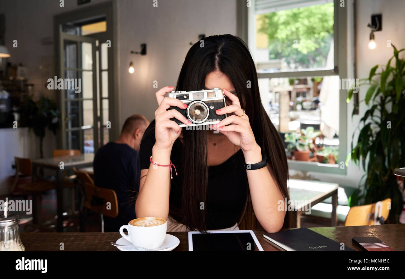 Brunette girl taking a picture with her film camera in a coffee shop next to the window. Stock Photo