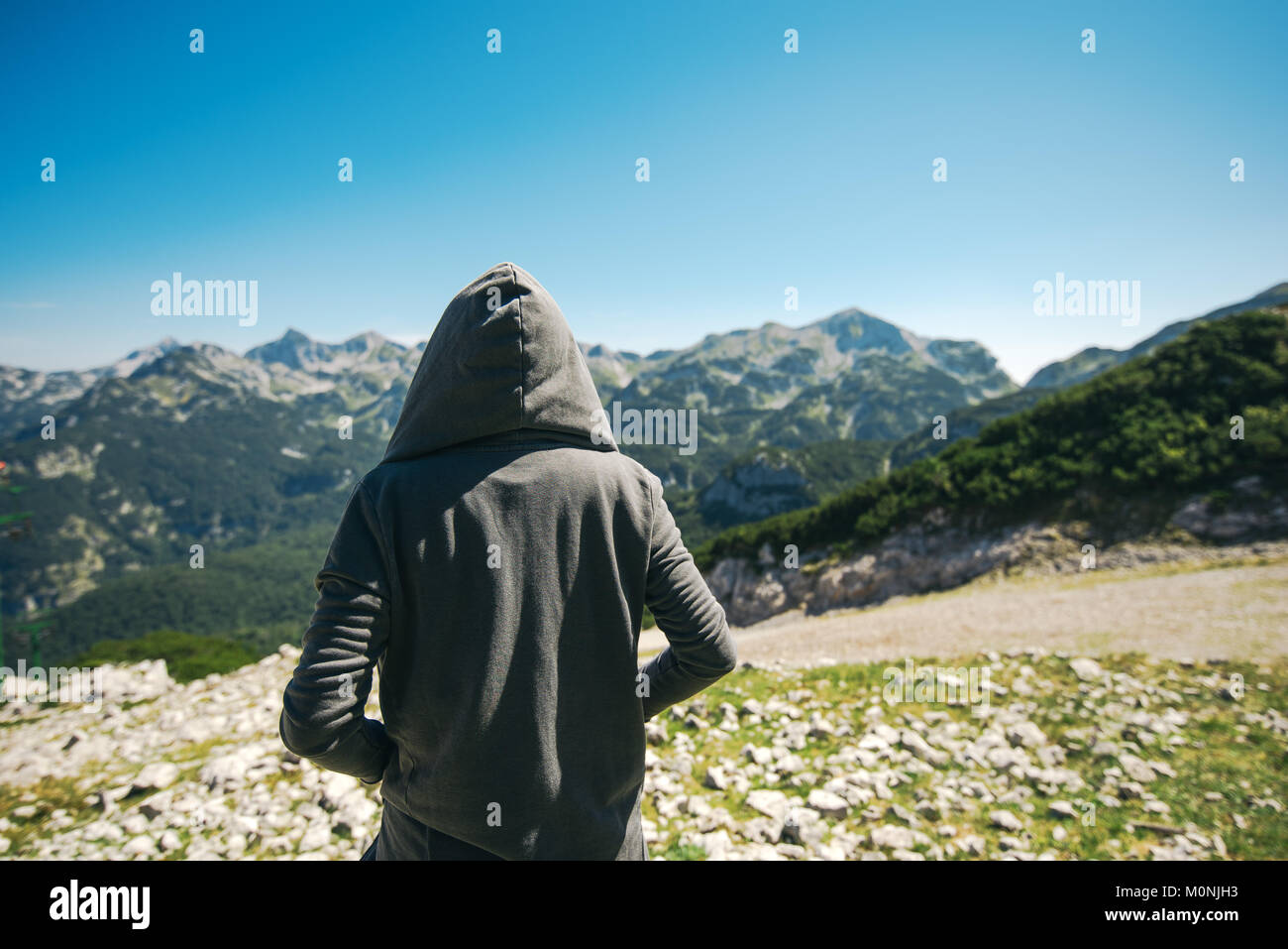 Mountain hiker at high viewpoint looking at the valley. Female tourist person in hooded jacket at mountain top enjoying the view. Stock Photo
