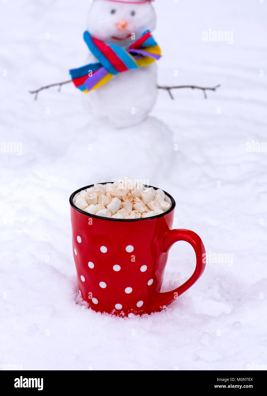 red ceramic mug with hot chocolate and marshmallow, behind a cheerful snowman in the snow Stock Photo