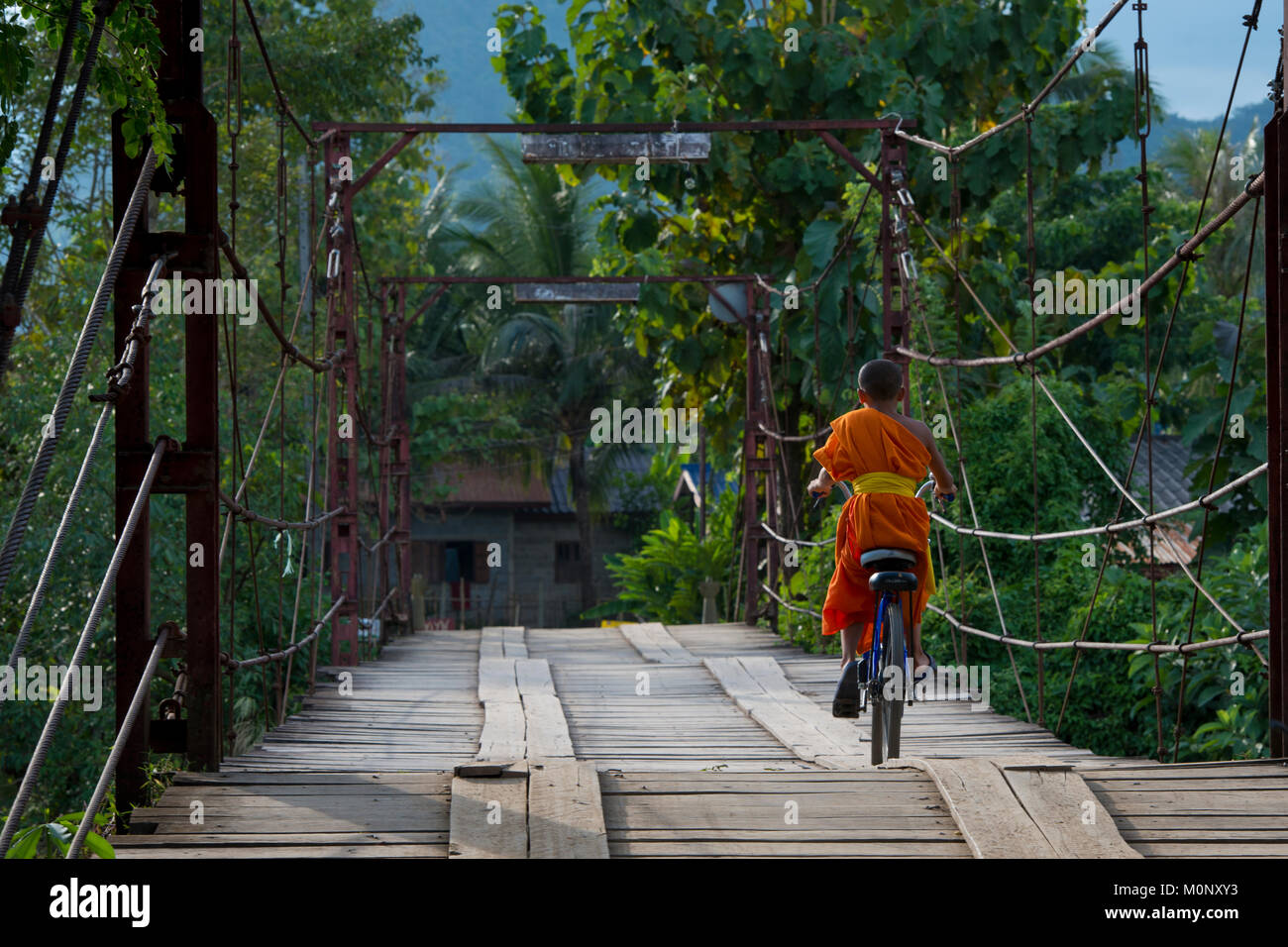 Novice,young monk,with bicycle on wooden bridge over the Nam Song River,Vang Vieng,Laos Stock Photo