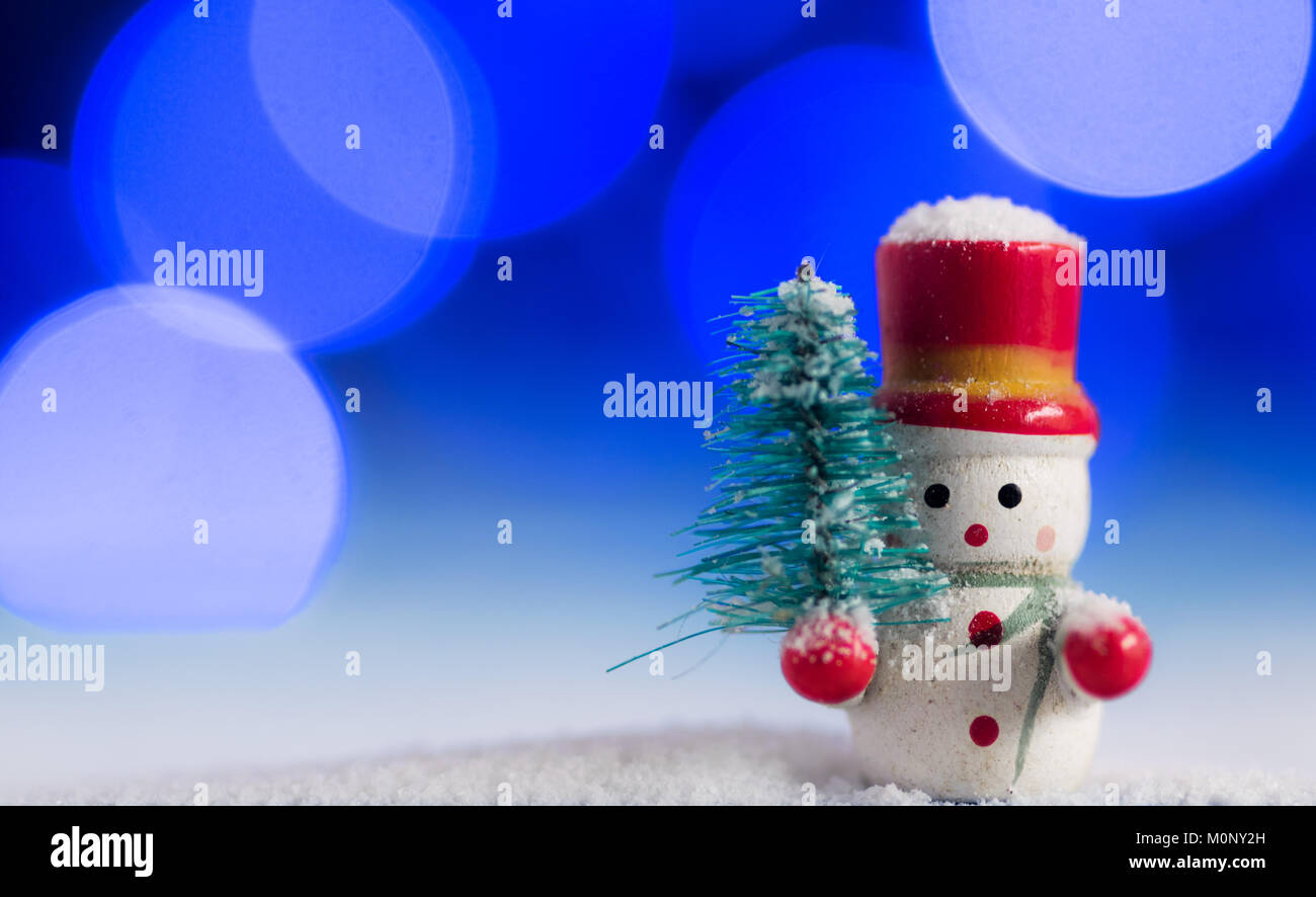 Cute Snowman christmas decoration, with beautiful blue and purple bokeh lights background, on top of white snow, holding a pine tree Stock Photo