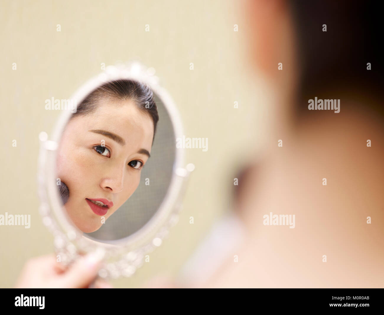 beautiful young asian woman looking at self in mirror while applying make-up on face using brush. Stock Photo