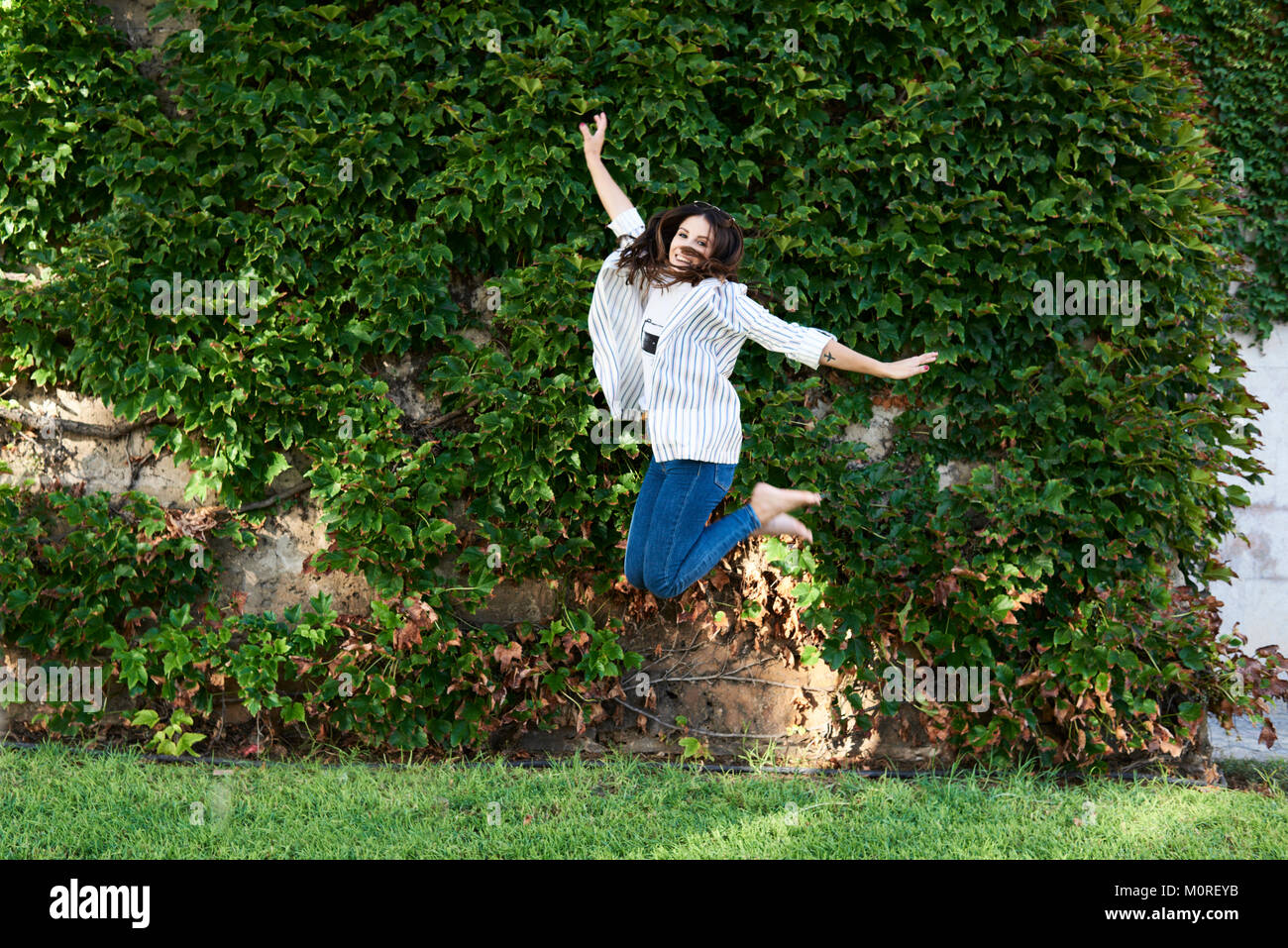 Happy millennial girl jumping freely against garden wall in summer. Stock Photo