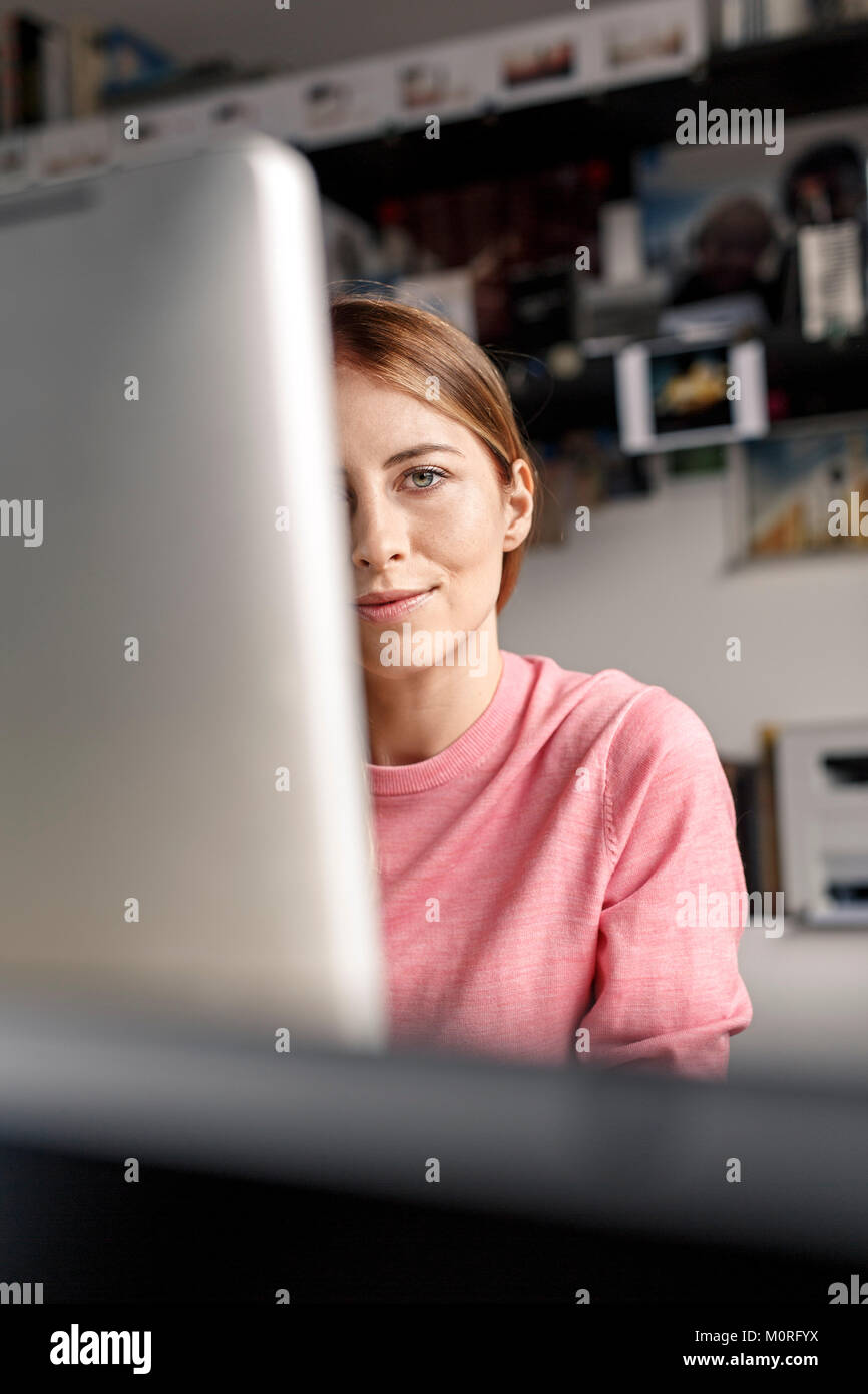 Portrait of young woman behind computer screen at desk at home Stock