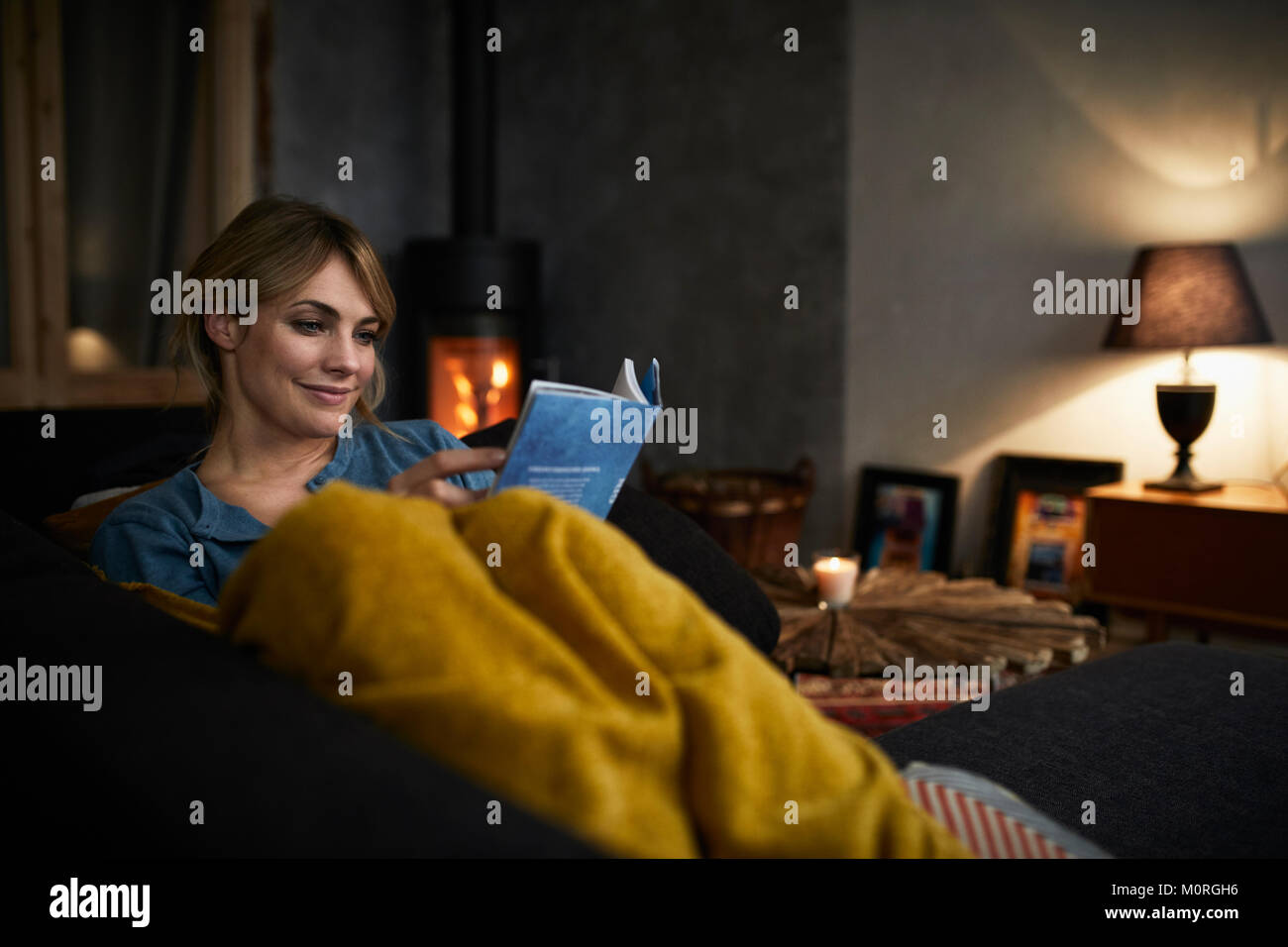 Portrait of smiling woman reading a book on couch at home in the evening Stock Photo