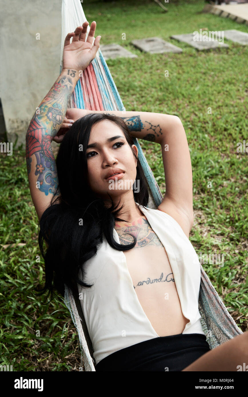 Tattooed asian female lying on a hammock relaxed and pensive in her summer villa. Stock Photo