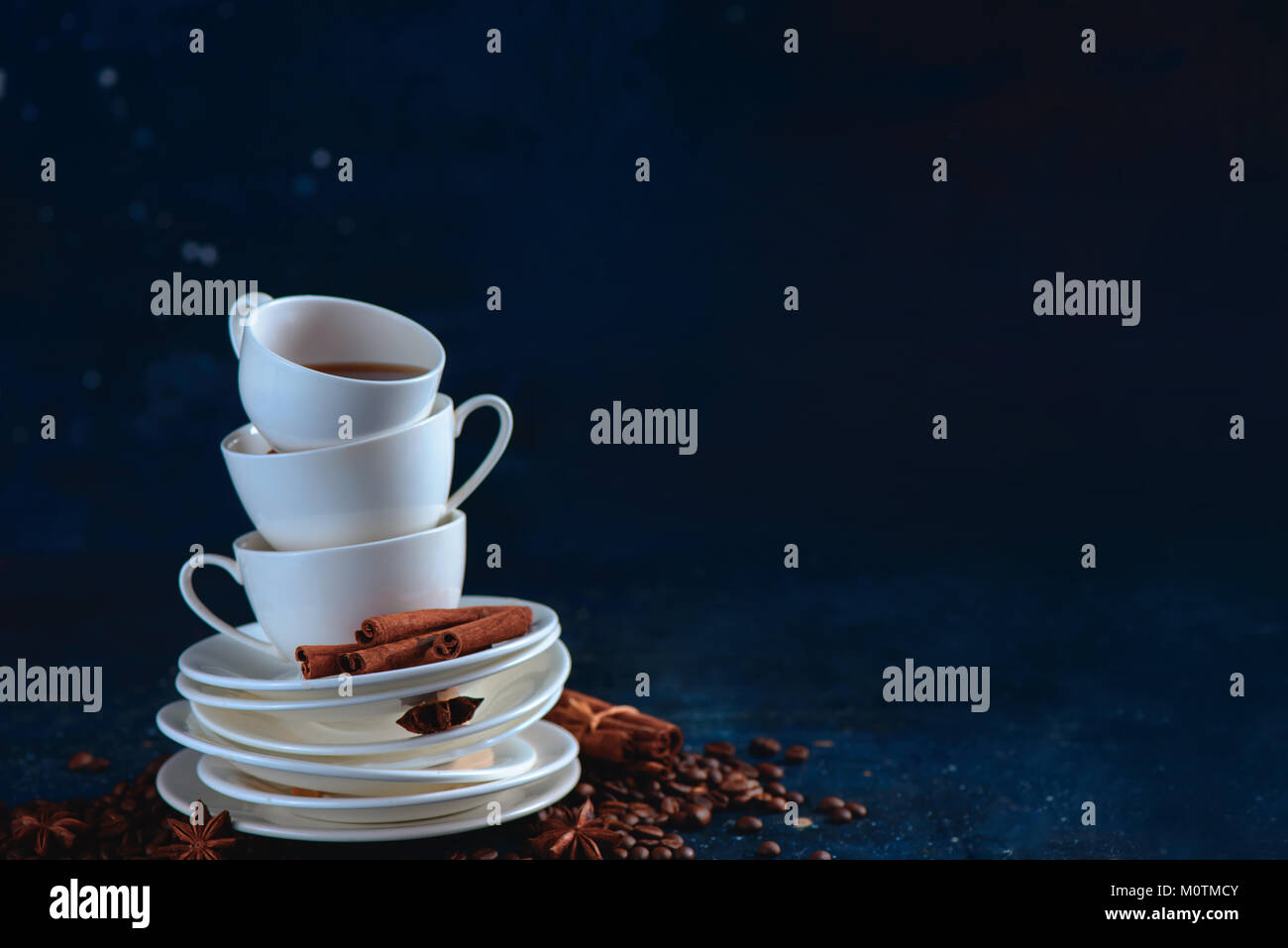 Header with a stack of balancing white coffee cups and tea saucers, coffee beans and cinnamon on a dark blue background. Dark food photography with co Stock Photo
