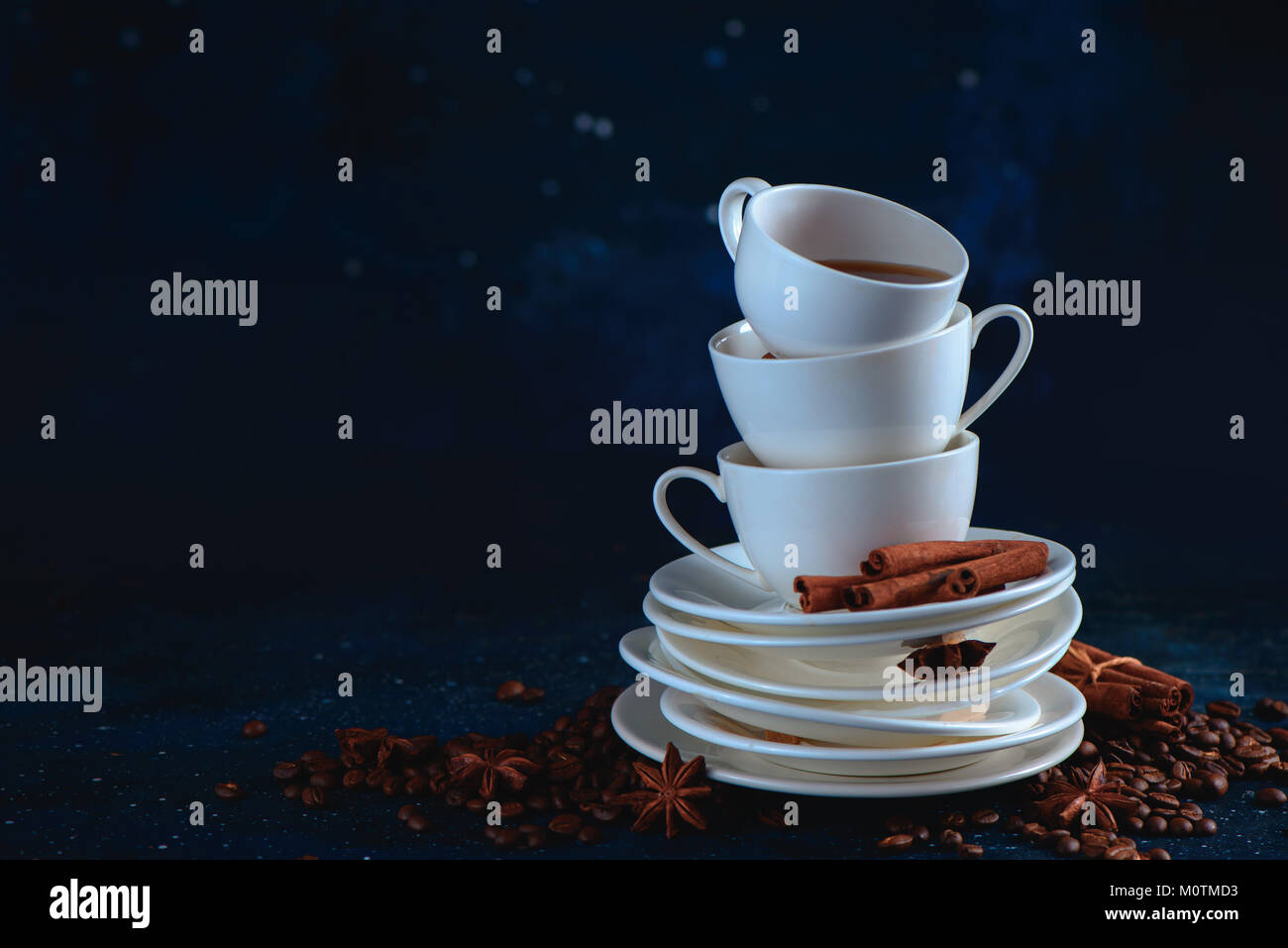 Header with a stack of balancing white coffee cups and tea saucers, coffee beans and cinnamon on a dark blue background. Dark food photography with co Stock Photo