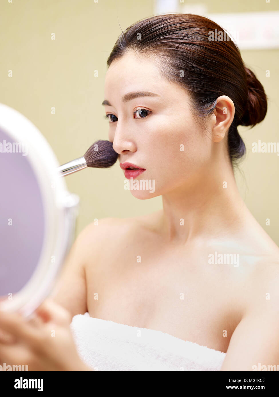 beautiful young asian woman looking in the mirror while applying make-up on face using brush. Stock Photo