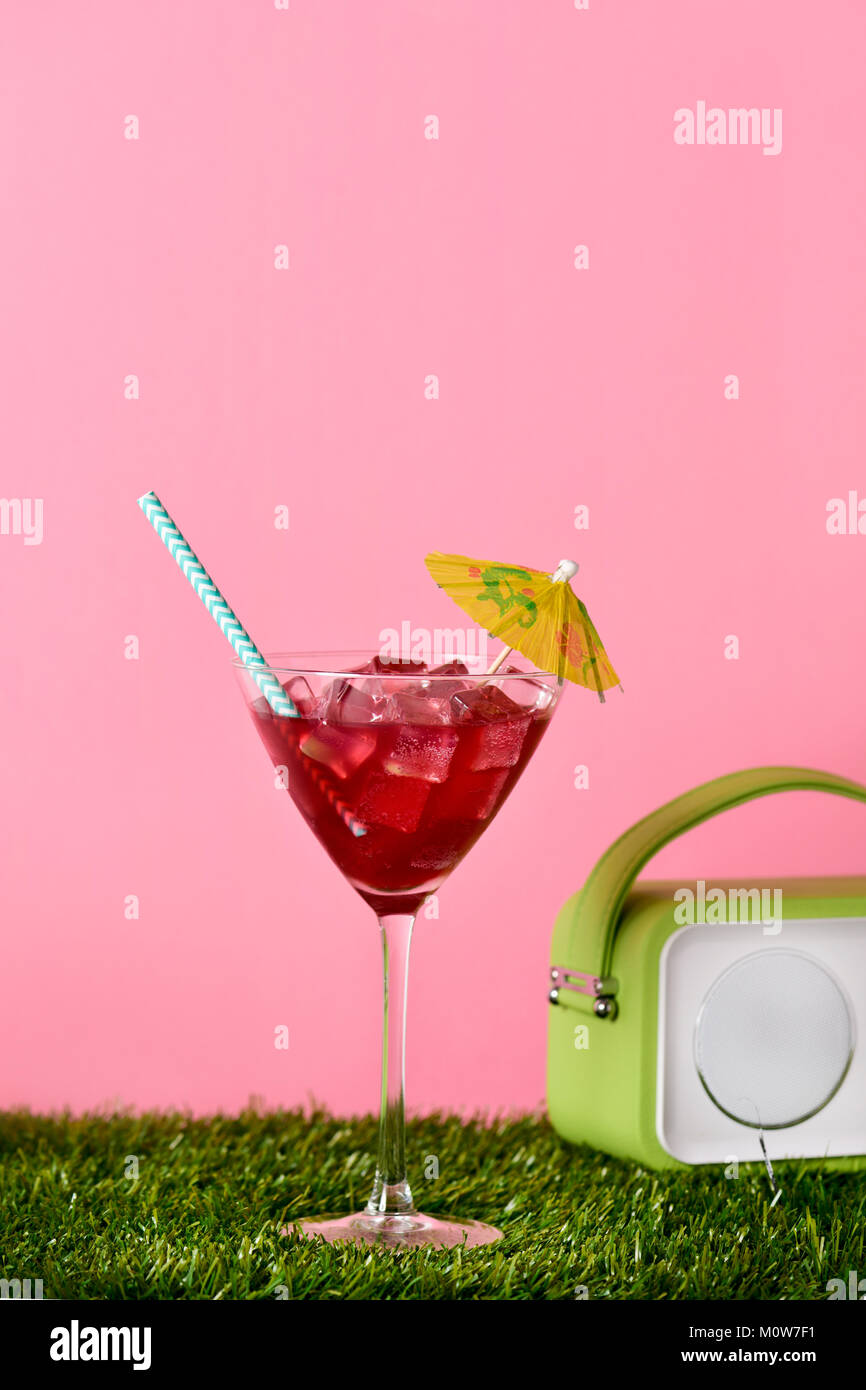 a cocktail glass with a red beverage garnished with a paper umbrella and a retro green radio on the turf, against a pink background with a blank space Stock Photo