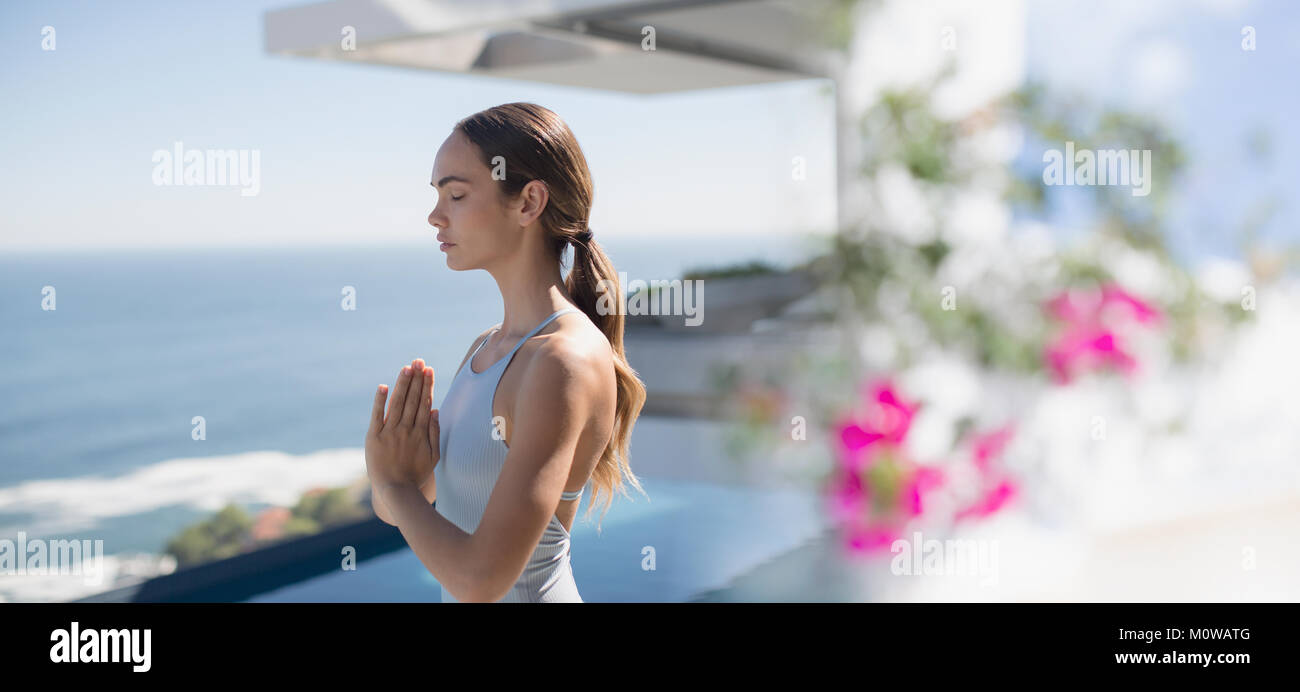Serene brunette woman practicing yoga, meditating with hands at heart center on sunny patio with ocean view Stock Photo