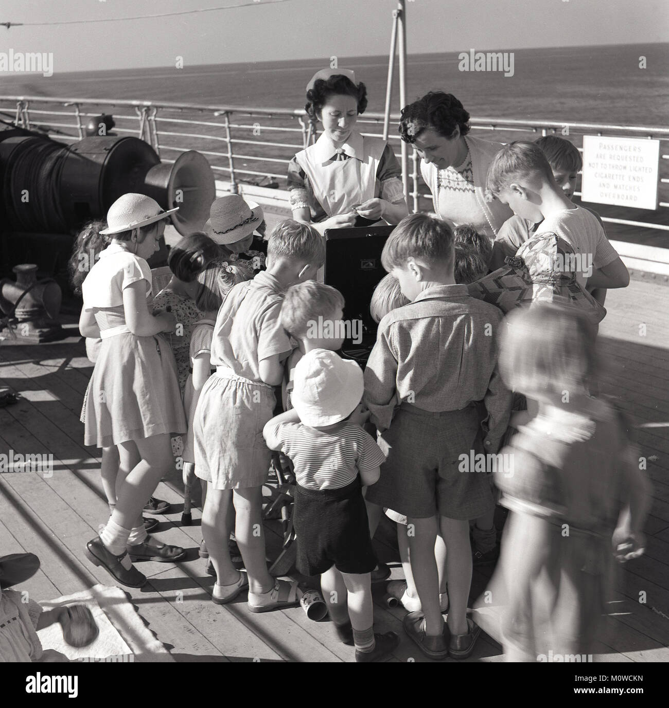 Mid 1950s, historical, aboard a Union-Castle steamship mid-ocean headed for the Cape in South Africa, taking mail, cargo and passengers, a group of children gather on the deck around a gramophone player, with one of the ship's nurse's and another lady supervising. Stock Photo