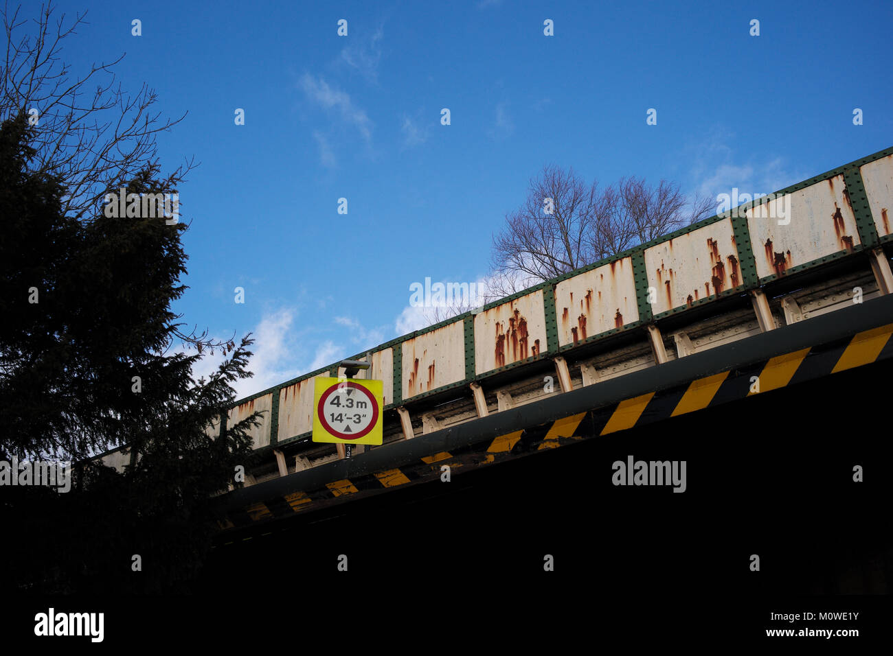 A close up of a rusty road crossing railway bridge with height warning signs against a blue sky. Stock Photo