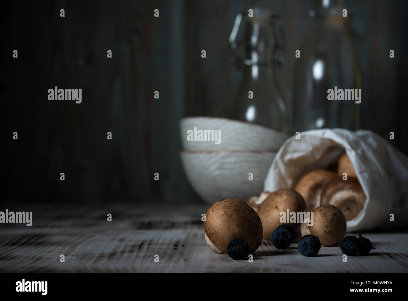 Horizontal photo of several brown edible mushrooms. Champignons are spilled on white wooden board from paper bag. Few dark blue backthorn berries are  Stock Photo