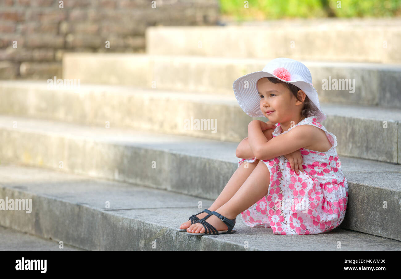 Adorable little girl wearing white hat  sitting on stairs on warm and sunny summer day Stock Photo