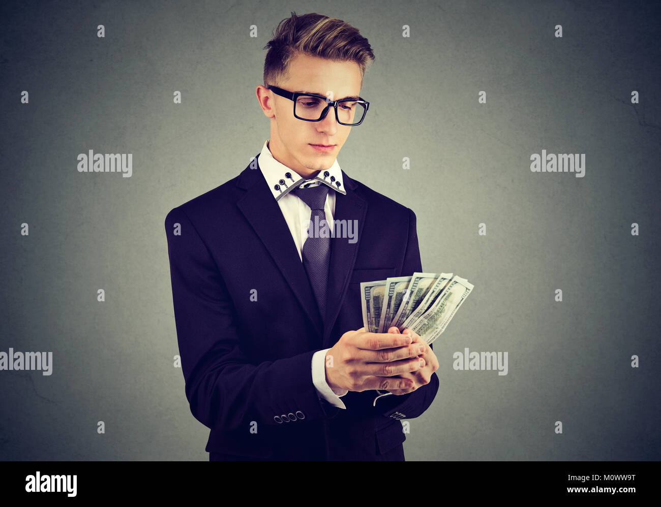 Young ambitious man in suit counting dollar bills in hands posing on gray. Stock Photo