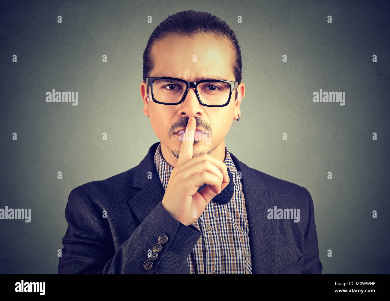 Young formal man in eyeglasses holding finger over lips asking for silence and looking at camera. Stock Photo