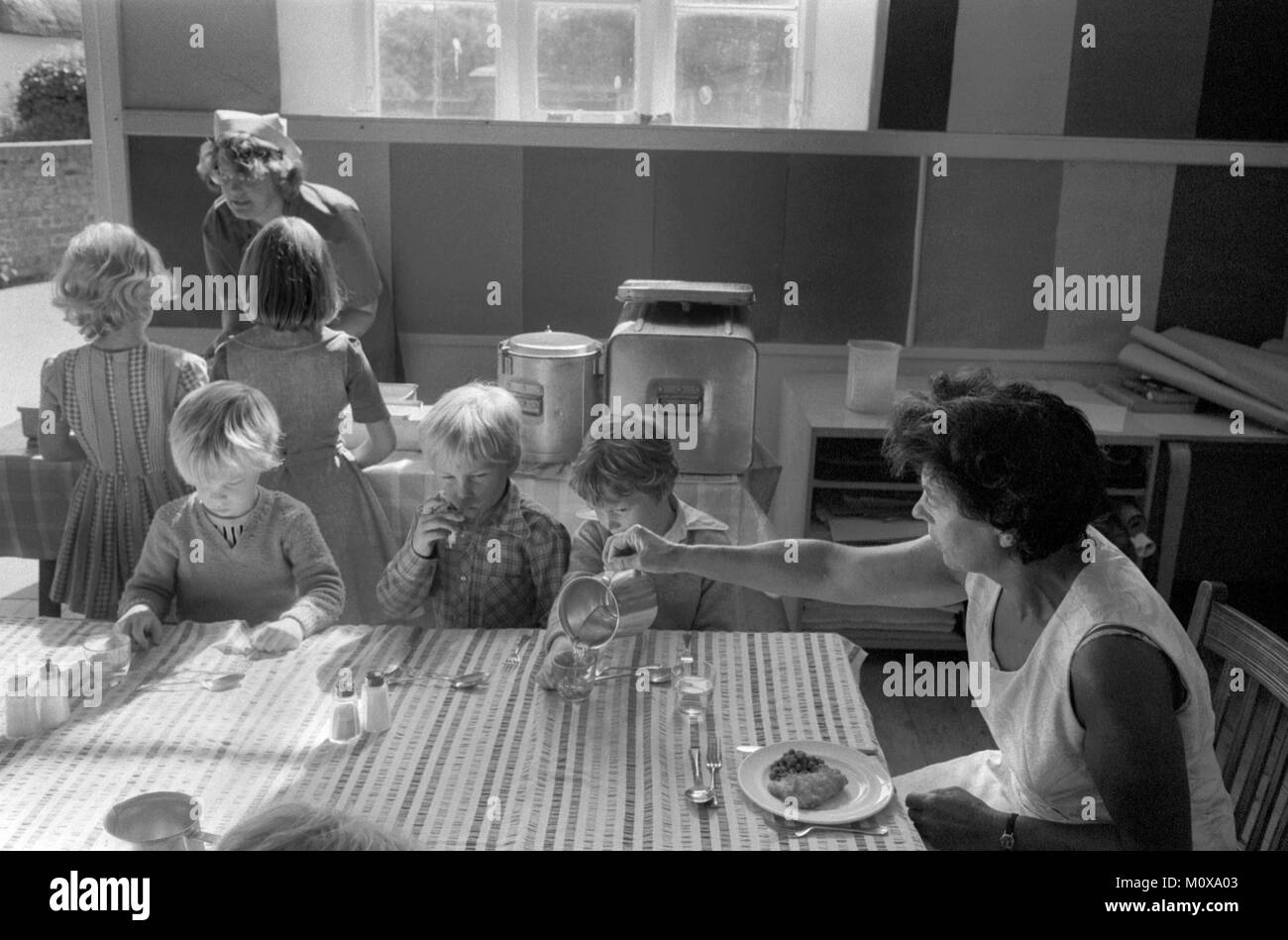 Village Primary school 1970s England. School children sit down together at one table with a member of staff and have lunch. School dinner lady. Cheveley Cambridgeshire 1978 UK HOMER SYKES Stock Photo