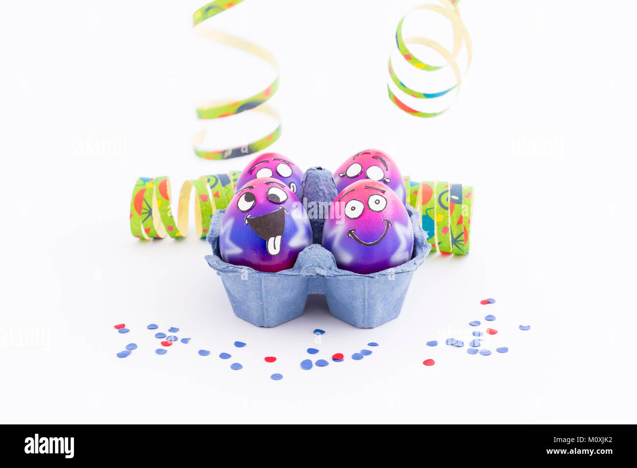 Group of colorful painted Easter eggs with funny cartoon style faces in a light blue egg box, colorful confetti and paper streamer on white background Stock Photo