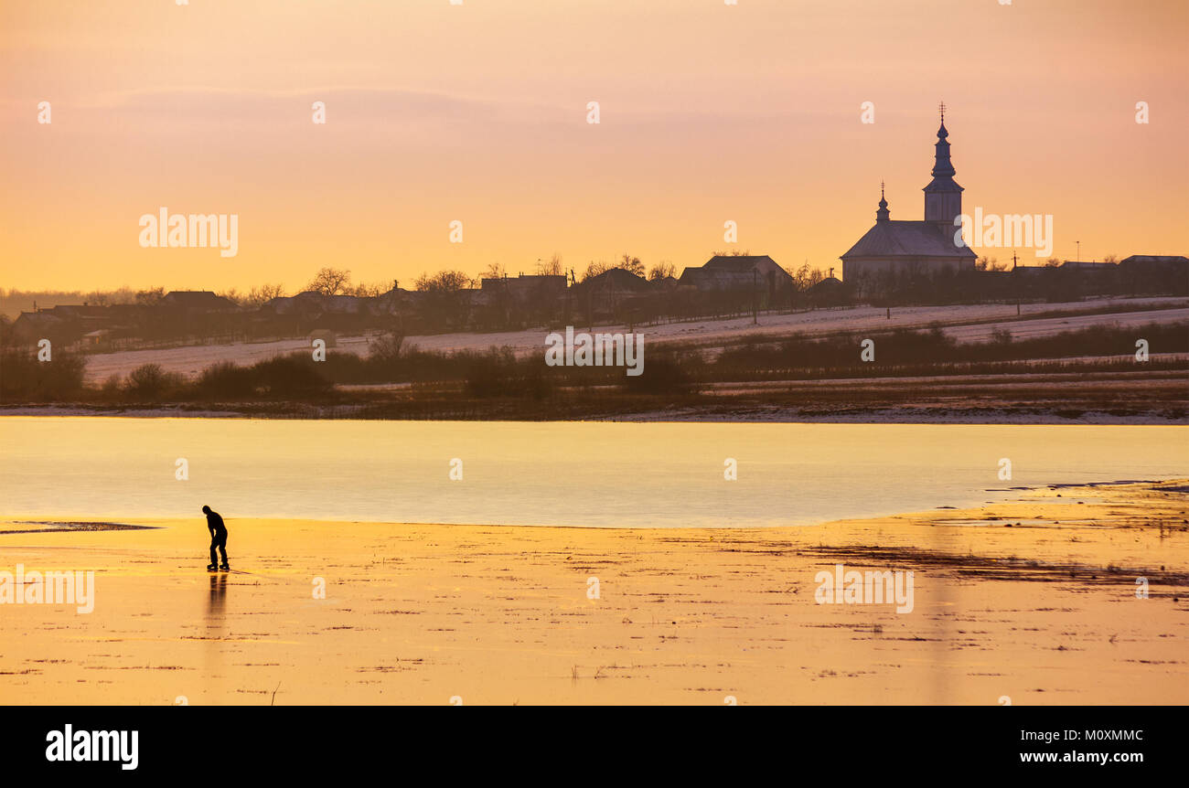 undefined person skating on the frozen lake in evening. beautiful winter countryside scenery. village and church in a far distance Stock Photo
