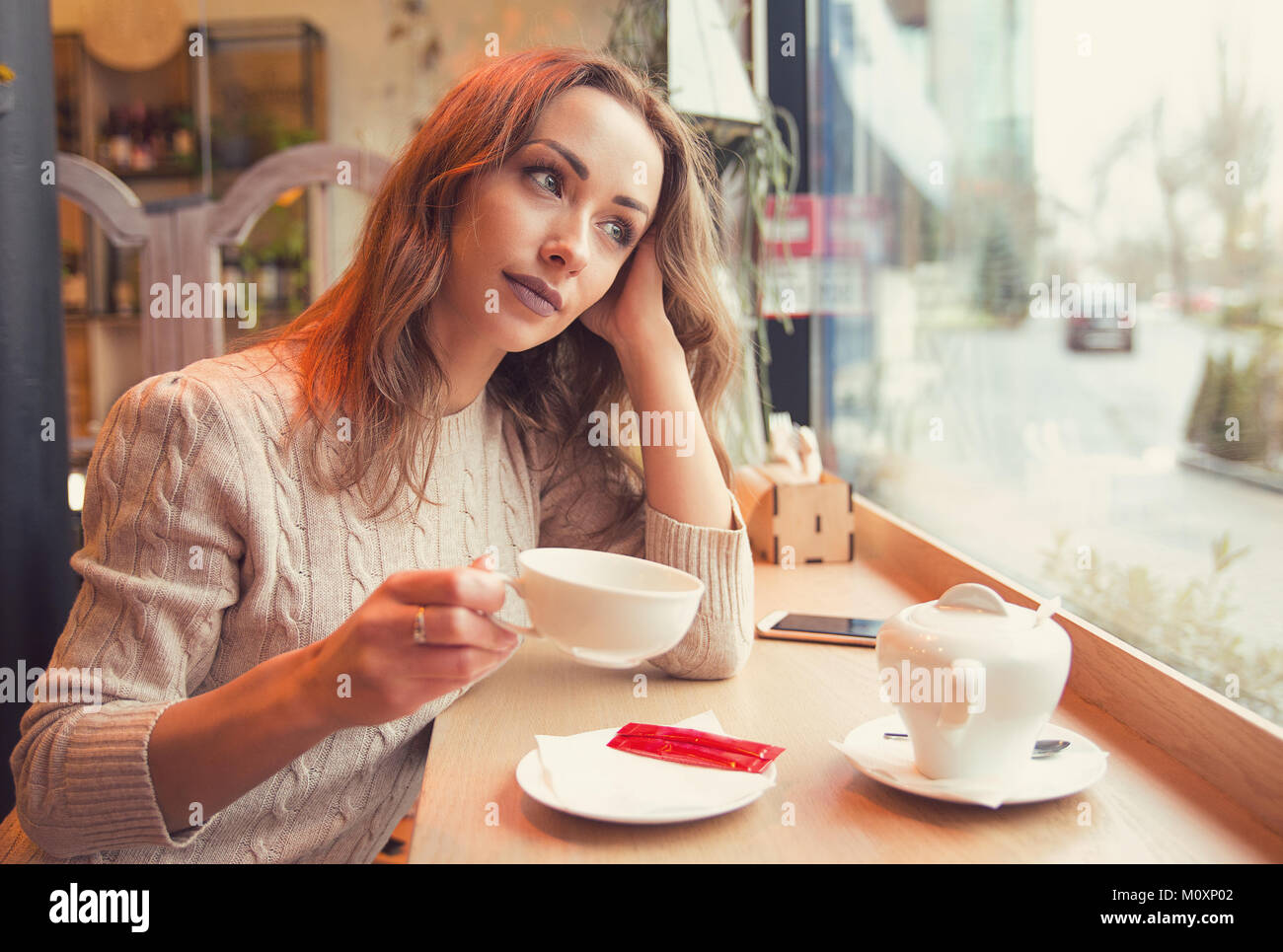 Young woman in sweater relaxing with cup of coffee in cozy cafeteria looking away and dreaming. Stock Photo