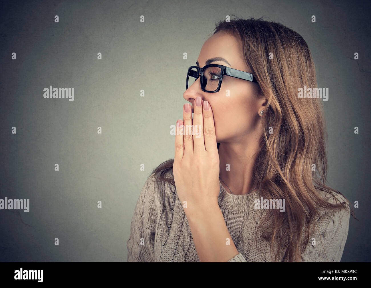Young woman covering mouth while telling secret posing on gray backdrop. Stock Photo
