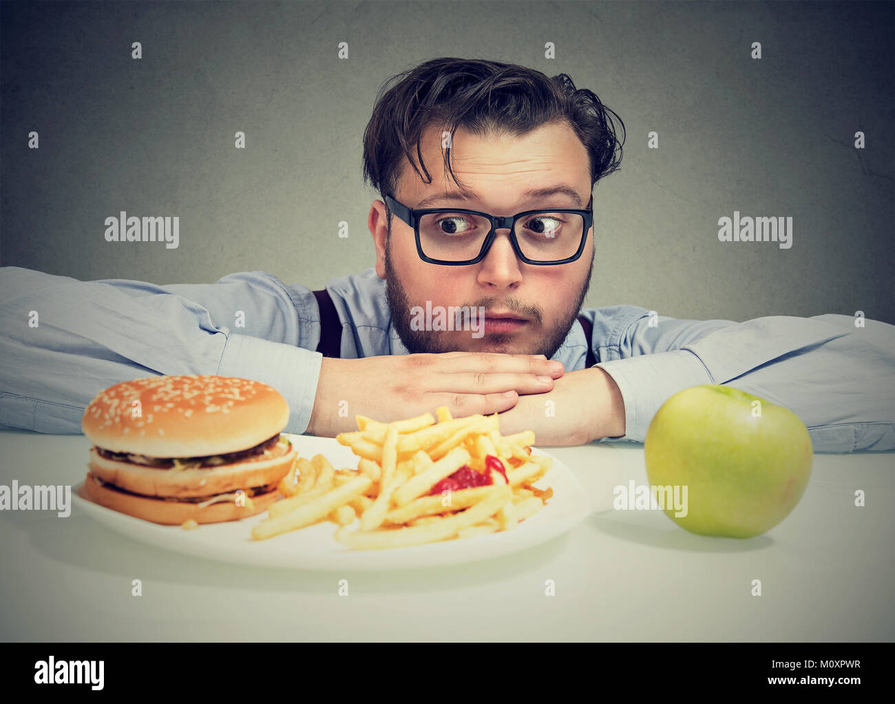 Young chunky man wanting tempting burger instead of healthy apple looking concerned. Stock Photo