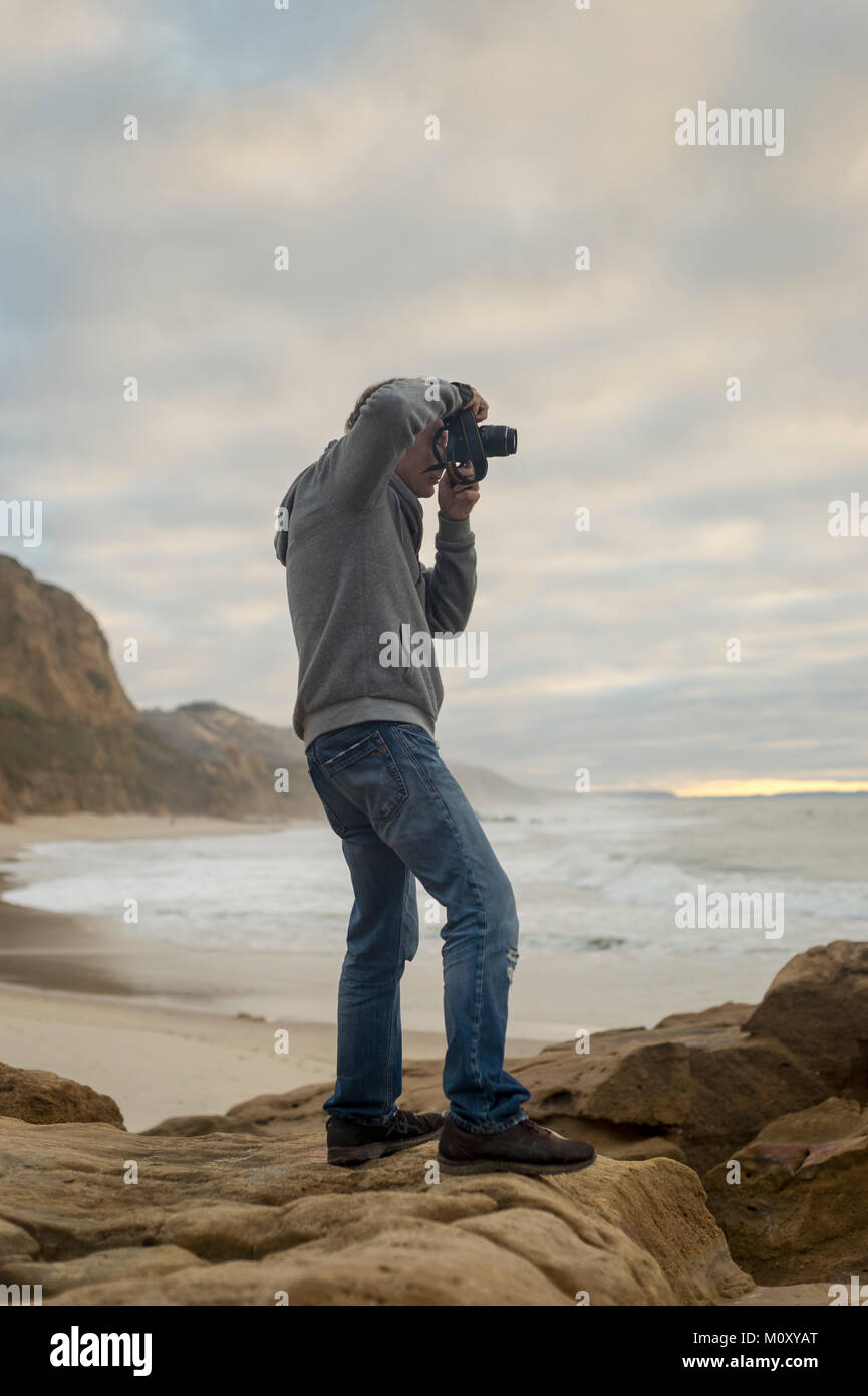 man standing on rocks by the coastline taking photographs with DSLR camera. Stock Photo