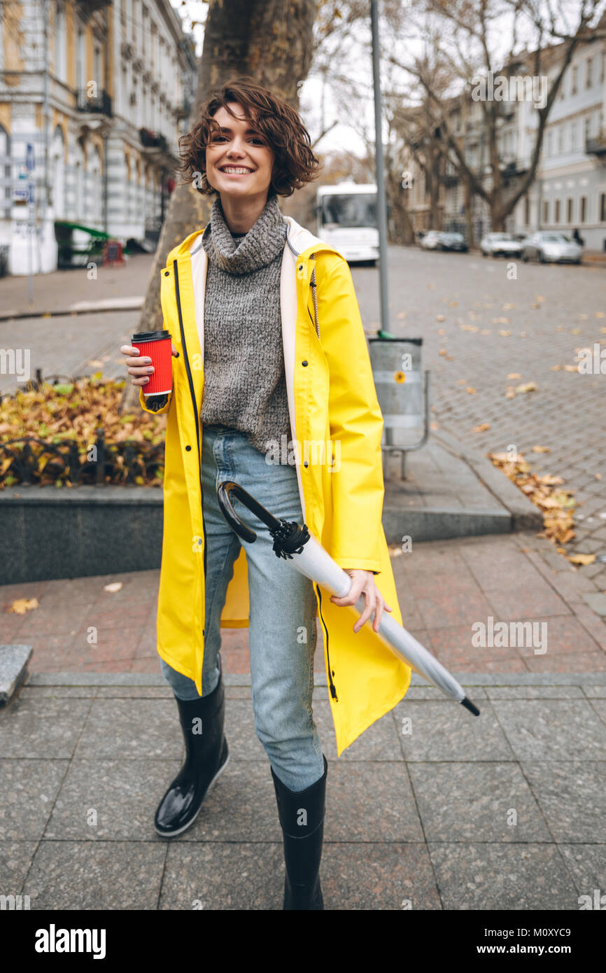 Photo of cheerful pretty young woman dressed in rubber boots and yellow  raincoat walking outdoors drinking coffee. Looking camera Stock Photo -  Alamy