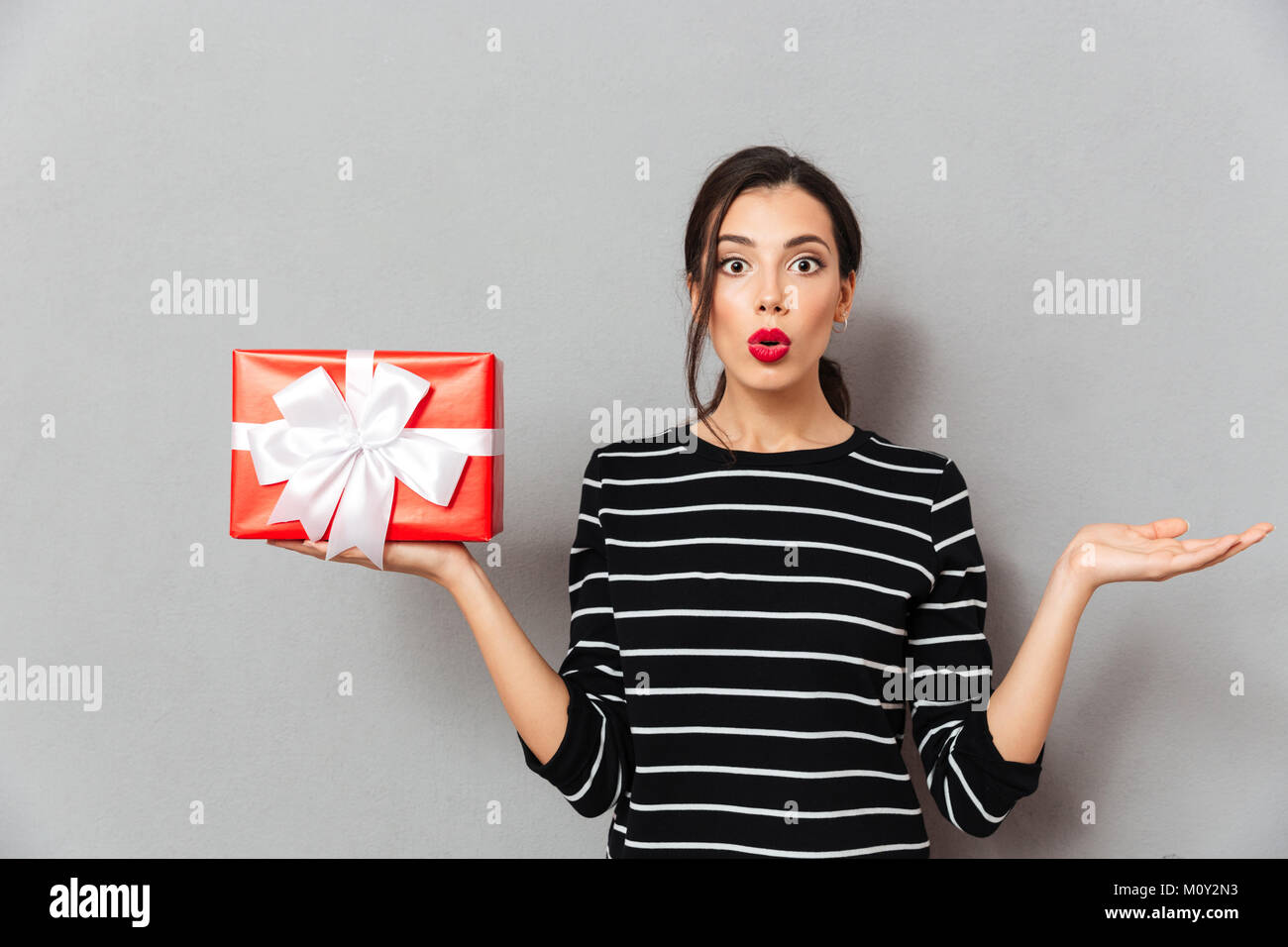 Portrait of a confused woman holding gift box and shrugging shoulders isolated over gray background Stock Photo