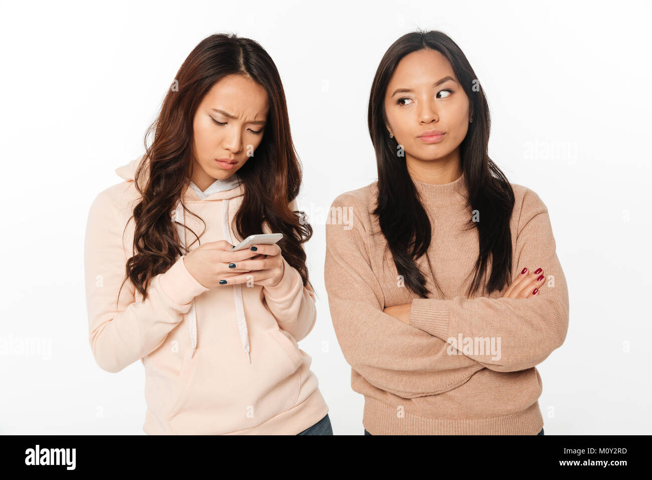 Photo of asian girl displeased because of her sister chatting by mobile phone. Looking aside. Stock Photo