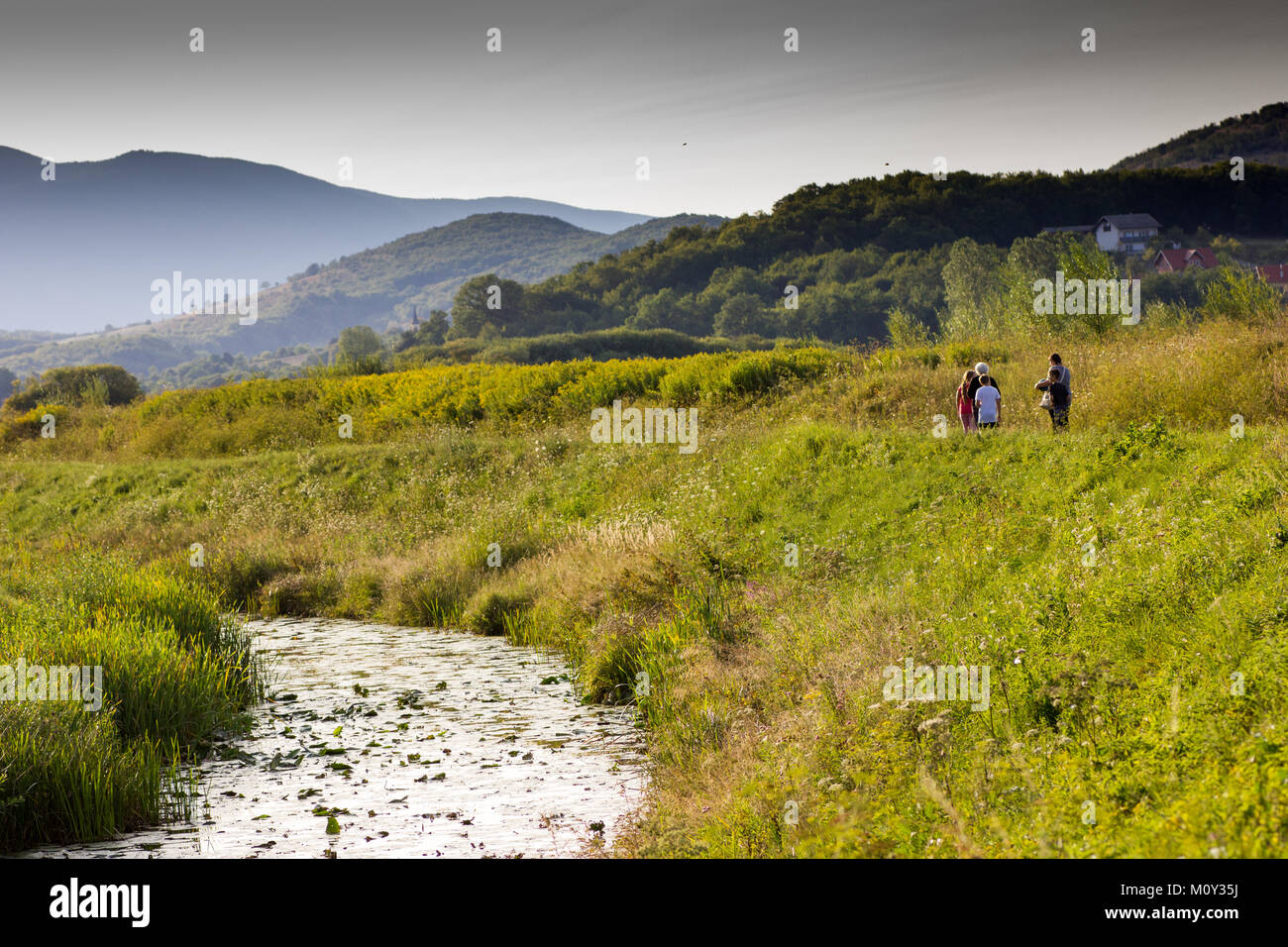 Old Gacka river passing through green mountainous countryside with small group of people (including children) walking along the grassy bank Stock Photo