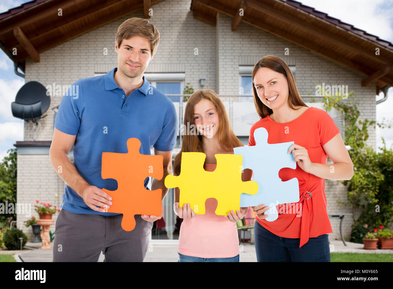 Portrait Of Happy Family Standing In Front Of Their House Holding Colorful Jigsaw Puzzles Stock Photo