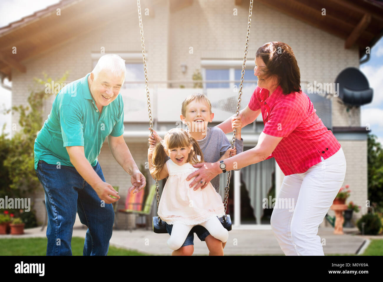 Happy Grandparents Looking At Their Grandchildren Playing On Swing In Front Of Their House Stock Photo