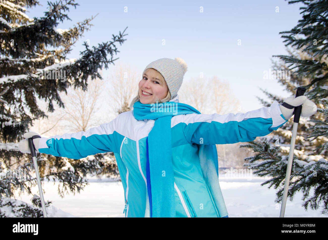 attractive smiling blonde with ski sticks in hands standing in winter forest, relaxing during skiing Stock Photo