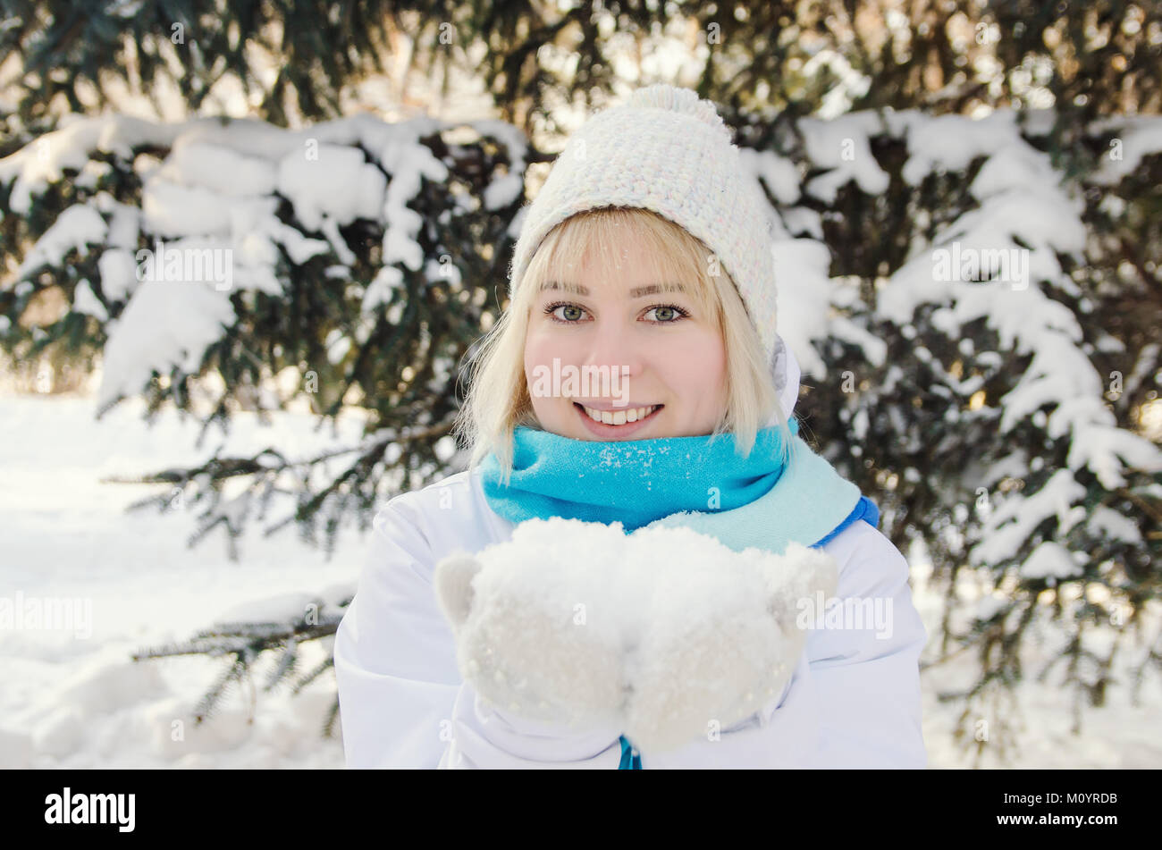 beautiful blond smiling girl holding snow on the palms against the background of winter snow-covered fir trees Stock Photo