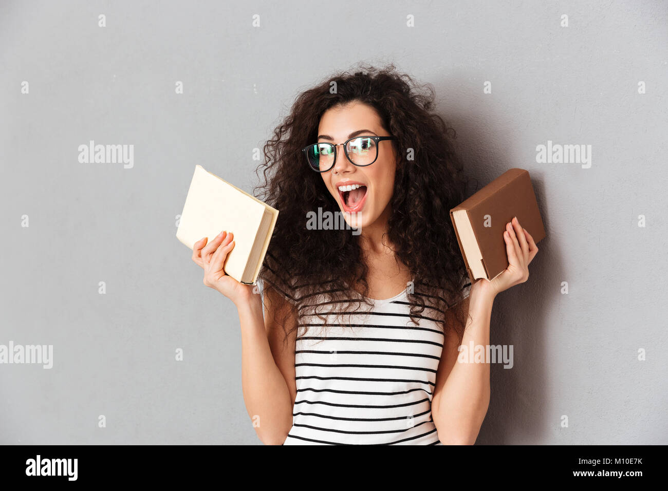 Woman with brown curly hair being student in university posing with interesting books in hands, taking pleasure in education isolated over grey wall Stock Photo