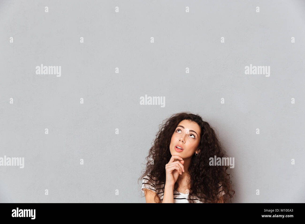 Concentrated young woman with shaggy hair touching her chin with face upward, and thinking or dreaming over grey background in studio Stock Photo