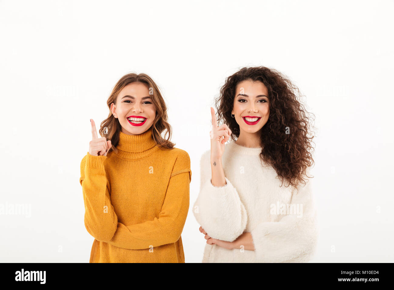 Two smiling girls in sweaters pointing up and looking at the camera over white background Stock Photo