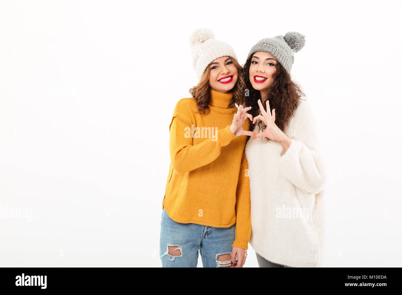Two smiling girls in sweaters and hats making heart sign while looking at the camera over white background Stock Photo