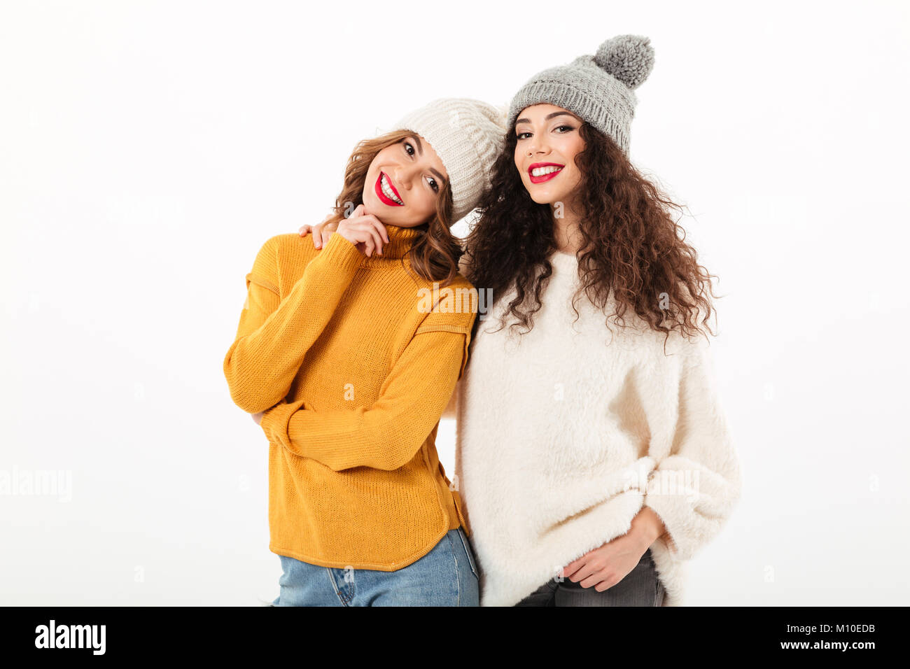 Two smiling girls in sweaters and hats posing together while looking at the camera over white background Stock Photo