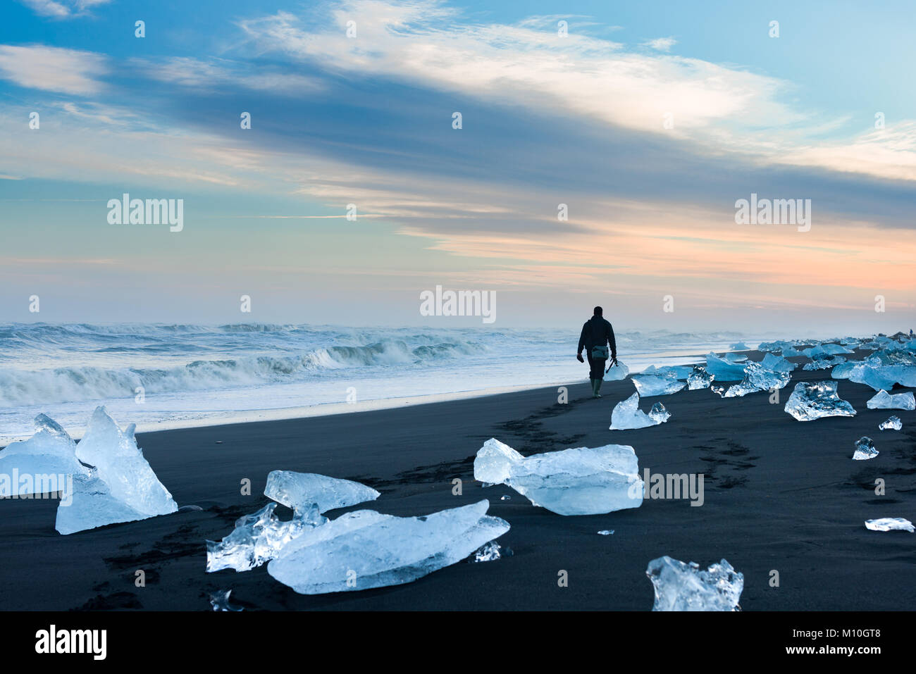 A male photographer walks amongst washed up icebergs along Jokulsarlon beach at sunset, Iceland Stock Photo