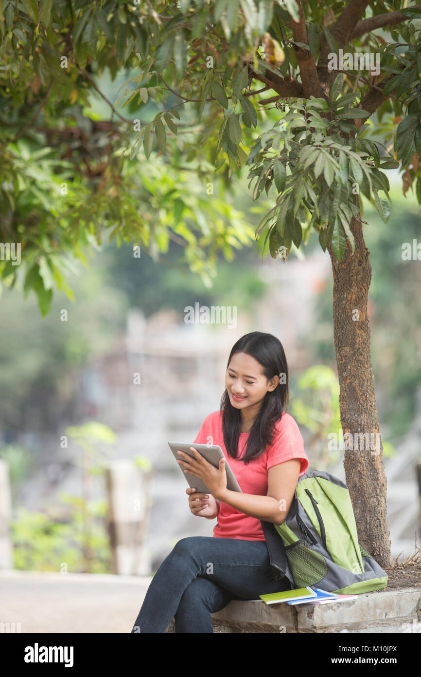 A portrait of a young Asian student sitting outdoor using tablet pc ...