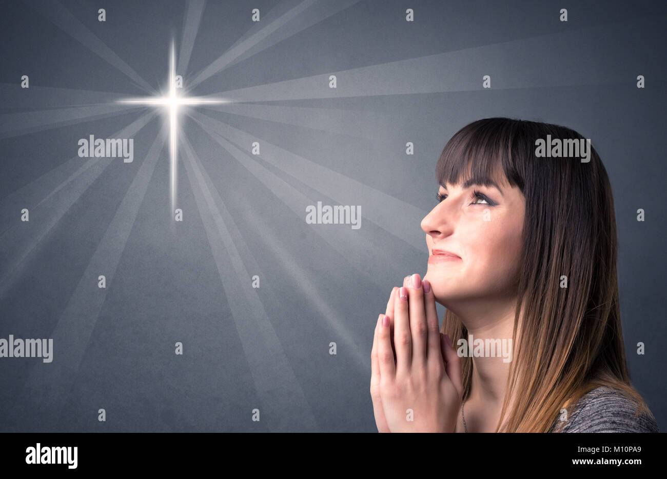 Young woman praying on a grey background with a shiny cross silhouette ...