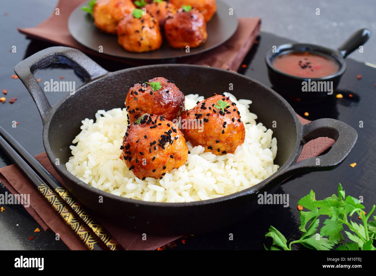 Spicy chicken balls in sweet chilli glaze with rice in a frying pan on a black wooden background Stock Photo