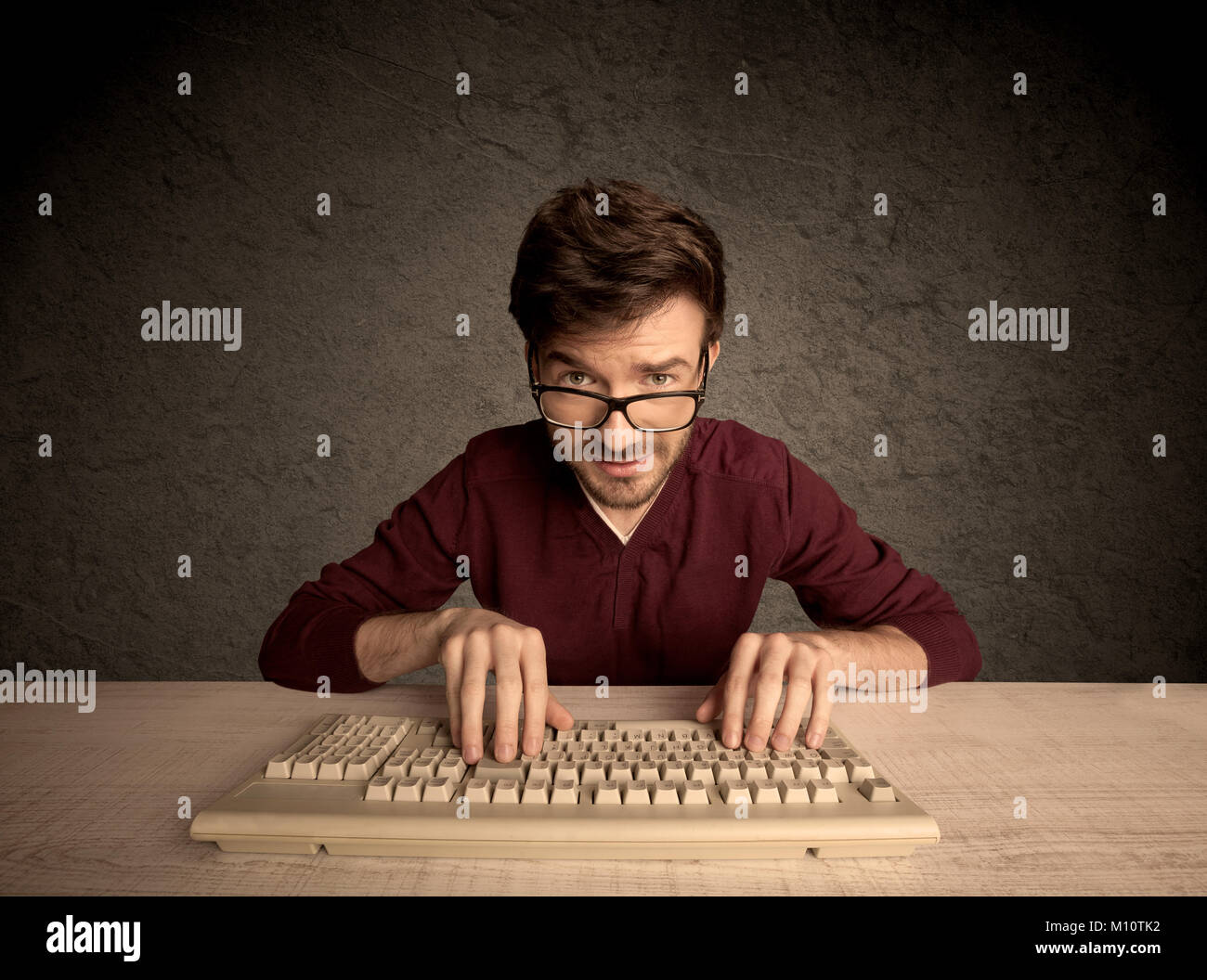 A young hacker with glasses dressed in casual clothes sitting at a desk and working on a computer keyboard in front of black clear concrete wall backg Stock Photo
