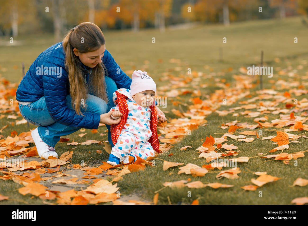Parent raises his daughter who fell making first steps. Baby girl learning to walk in autumn park. Walking toddler. Stock Photo