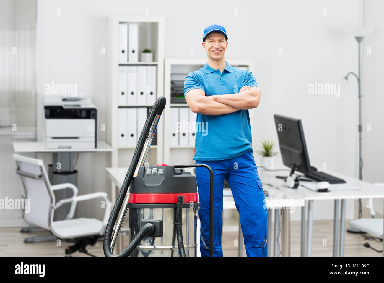 Portrait Of A Smiling Young Male Cleaner With Arms Crossed Standing In Office Stock Photo