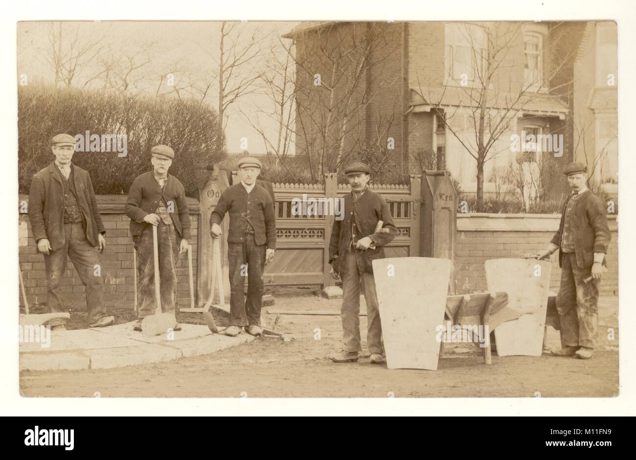 Original postcard of gang of Edwardian workmen, navvies, builders outside house, wearing working clothes and flat caps, holding their work tools, working on a pavement, road building, circa 1910, U.K. Stock Photo