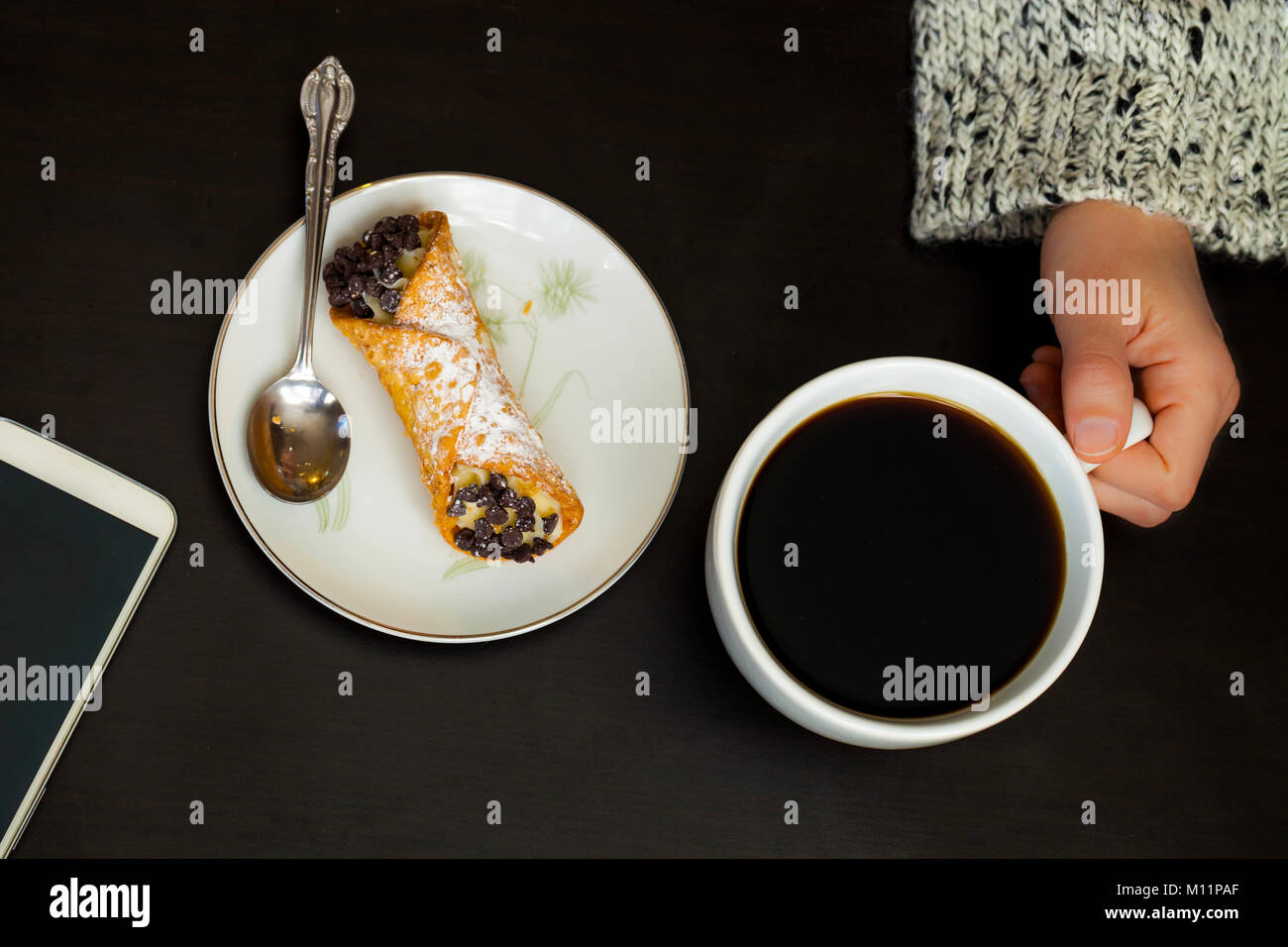 Caucasian woman hand holds coffee cup with smartphone and italian cannoli on plate on black background Stock Photo